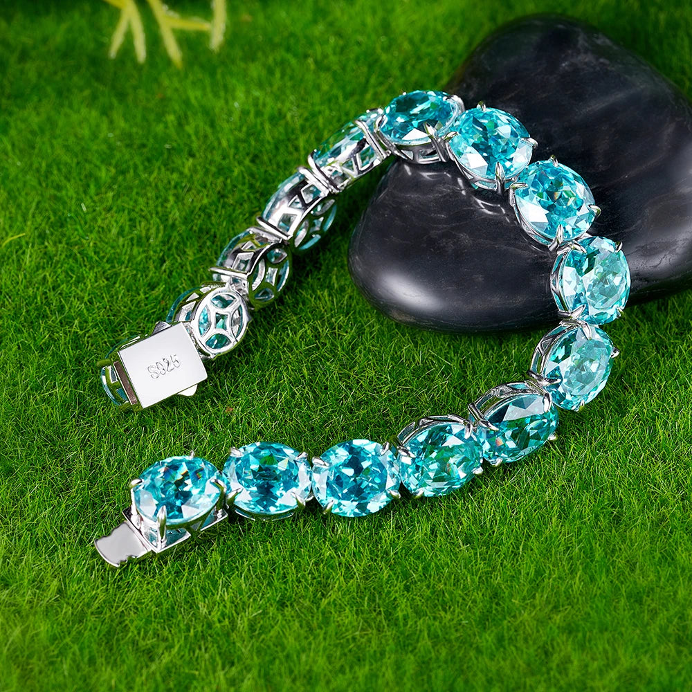 A silver-tone tennis bracelet with large, round, light blue gemstones, displayed on a green grass-like surface next to a black stone.