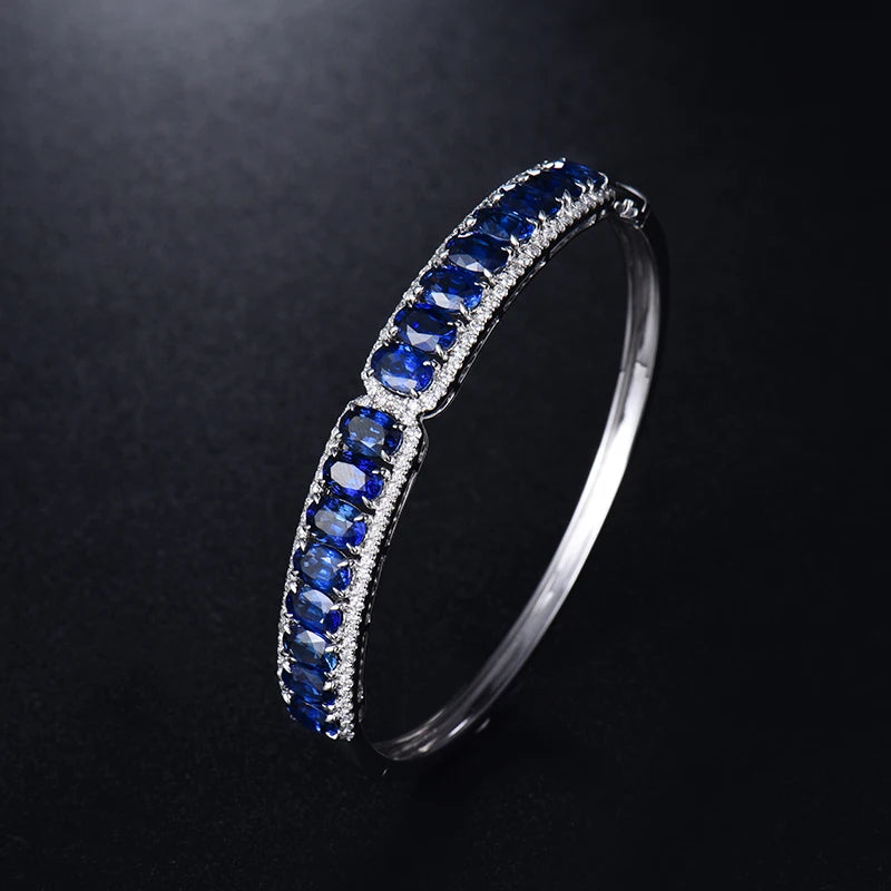 A silver-toned bangle bracelet featuring rows of deep blue oval-cut gemstones and small clear accent stones, photographed on a dark black background.