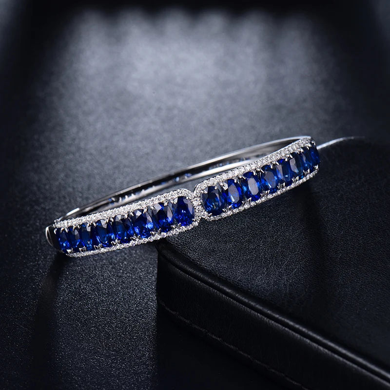 A silver-toned bangle bracelet featuring rows of deep blue oval gemstones and small clear accent stones, displayed on a dark textured surface.