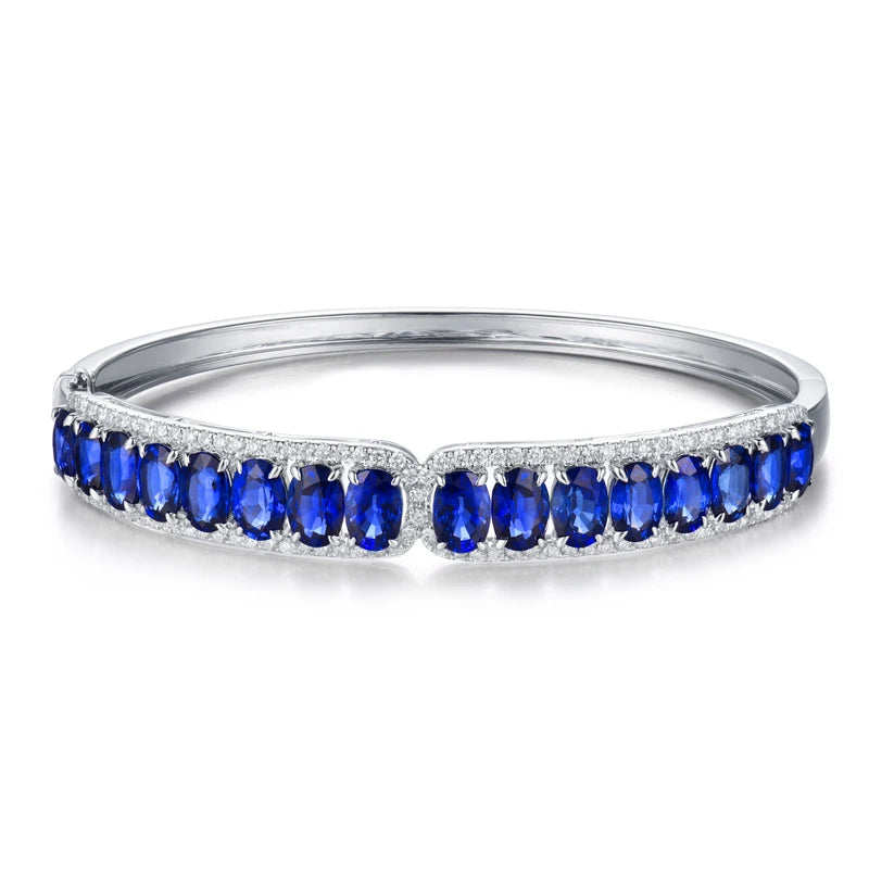 A silver-toned hinged bangle bracelet featuring a row of oval-cut deep blue sapphires and small clear accent stones, shown on a clean white background.