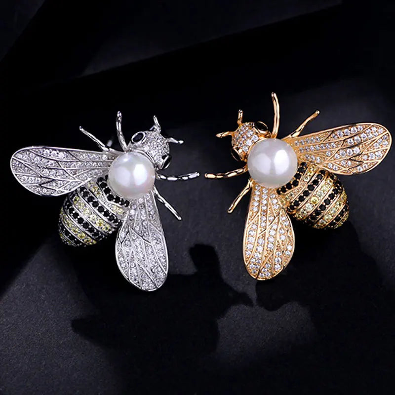Two bee-shaped brooches, one silver and one gold, with pearl centers on a black background.