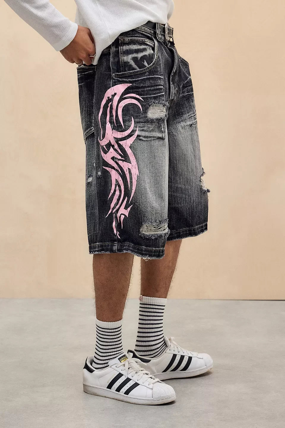 Person wearing black denim shorts with pink tribal design, white sneakers, and striped socks on a beige background.