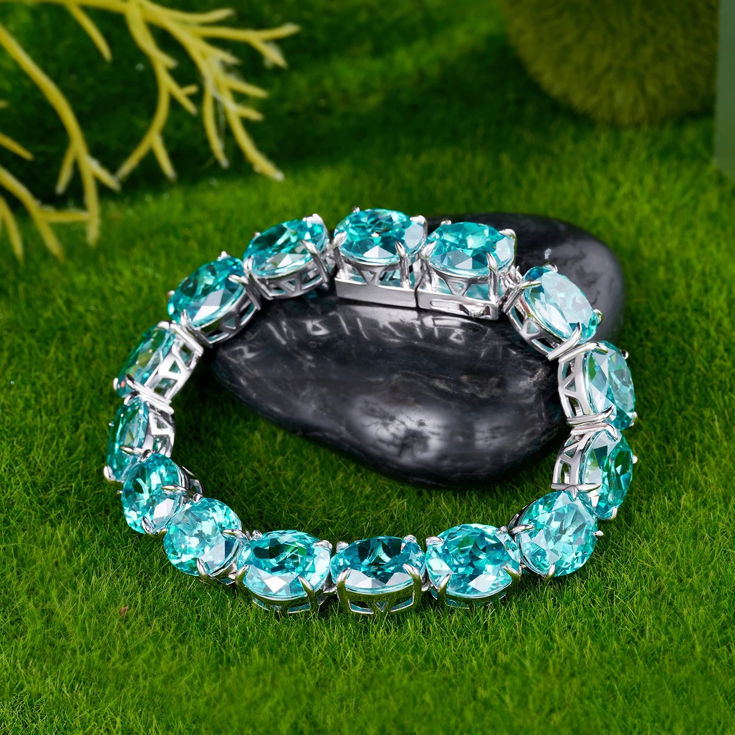 A silver-toned cuff bracelet set with large, round-cut light blue gemstones, displayed on a dark stone over a green grass-like surface.