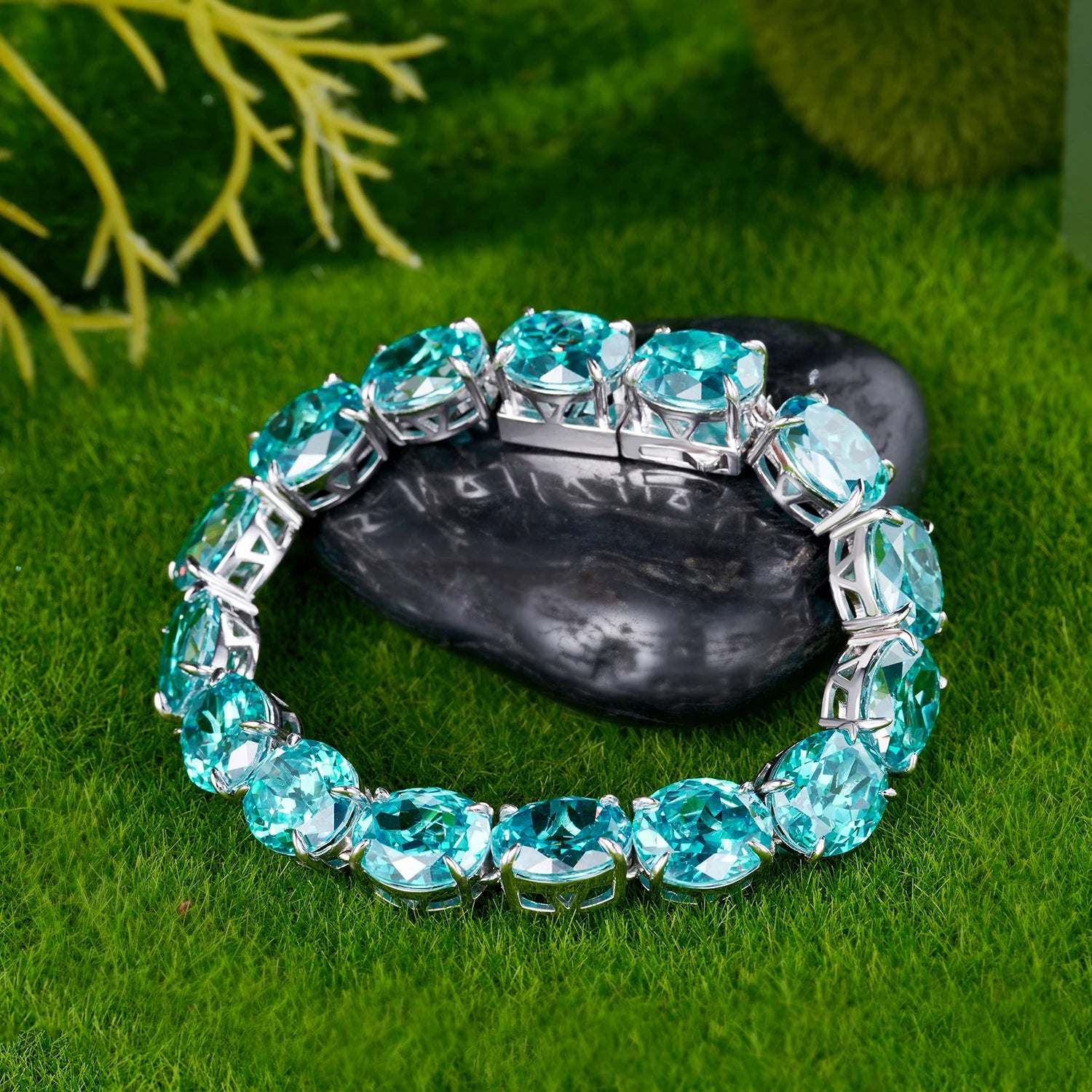A silver-toned cuff bracelet set with large, round-cut light blue gemstones, displayed on a dark stone over a green grass-like surface.