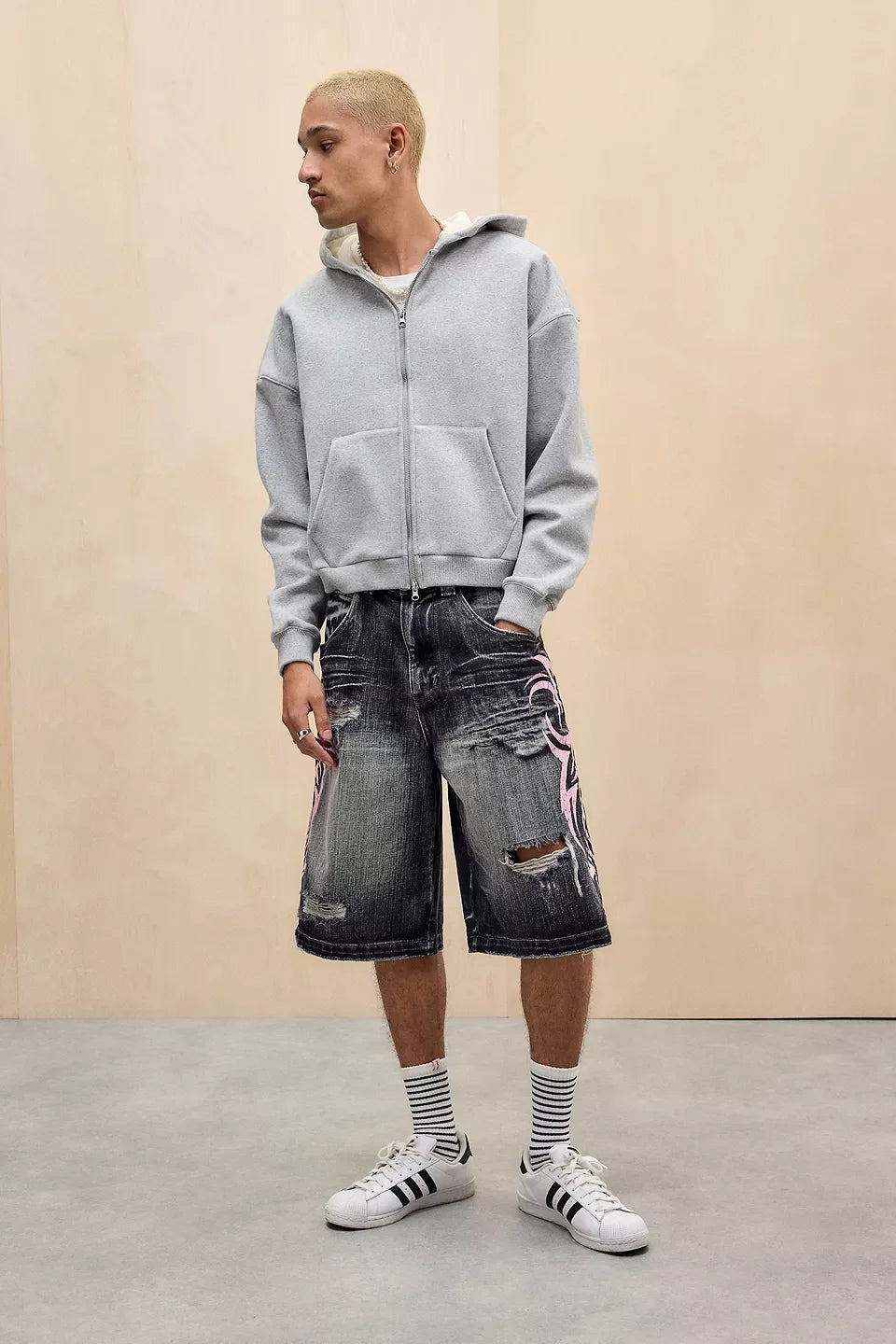 Person wearing a gray hoodie, black shorts with colorful designs, and white sneakers on a beige background