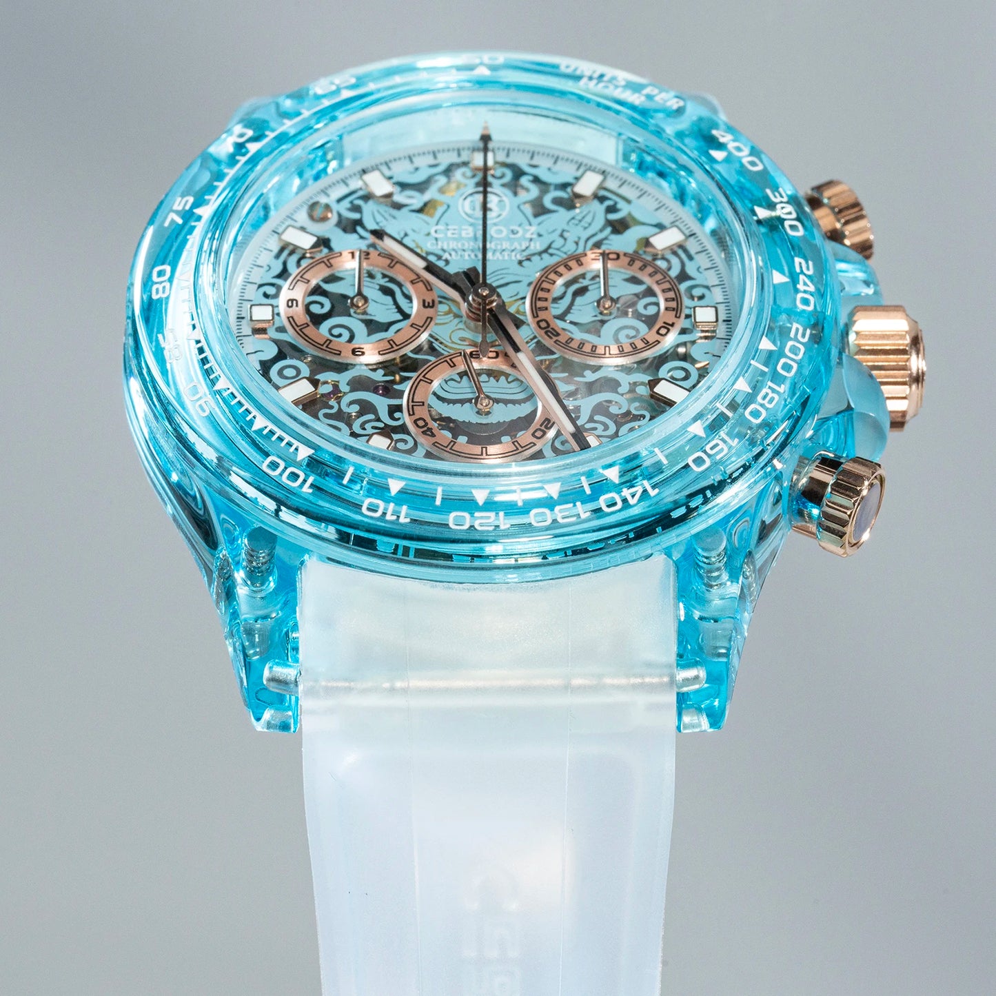A close-up of a Swatch wristwatch featuring a translucent light blue case, a white silicone strap, and rose gold-tone chronograph pushers and subdials.