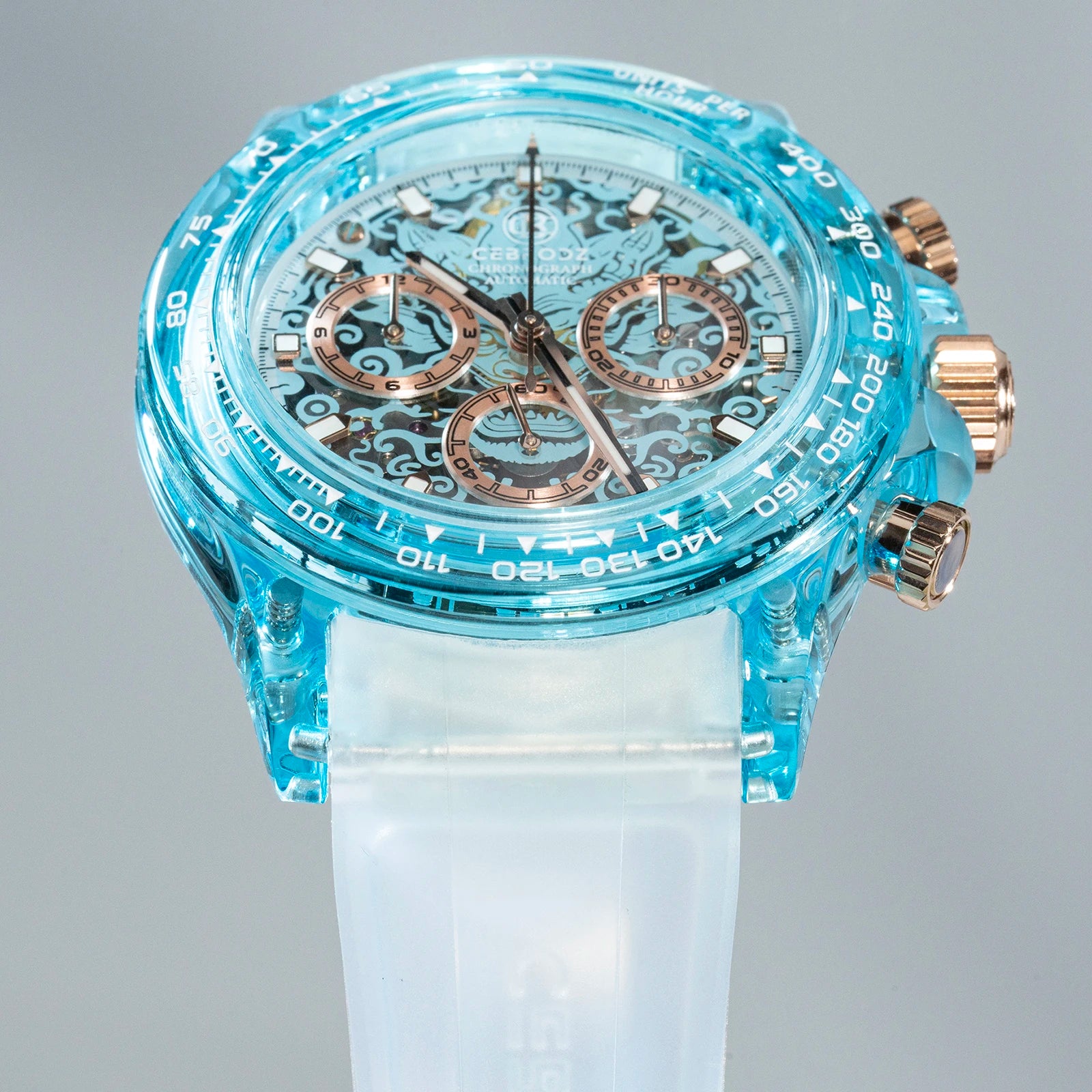 A close-up of a Swatch wristwatch featuring a translucent light blue case, a white silicone strap, and rose gold-tone chronograph pushers and subdials.