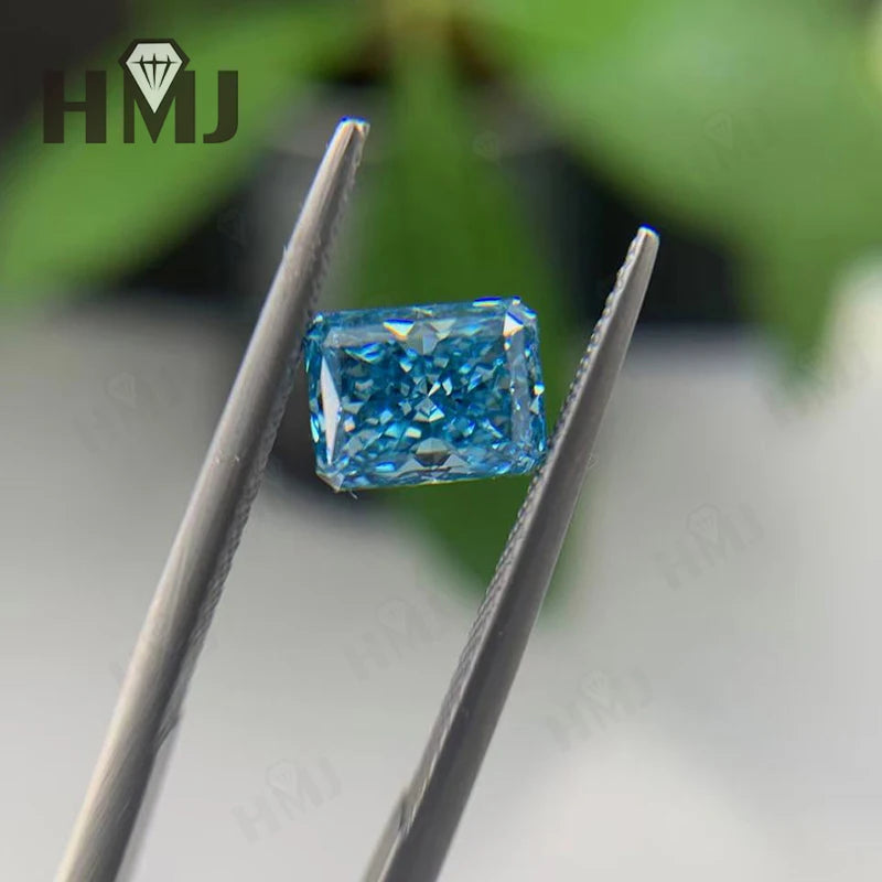 A rectangular blue gemstone held between metal tweezers, with a blurred green and gray background and an HMJ logo watermark in the top left.