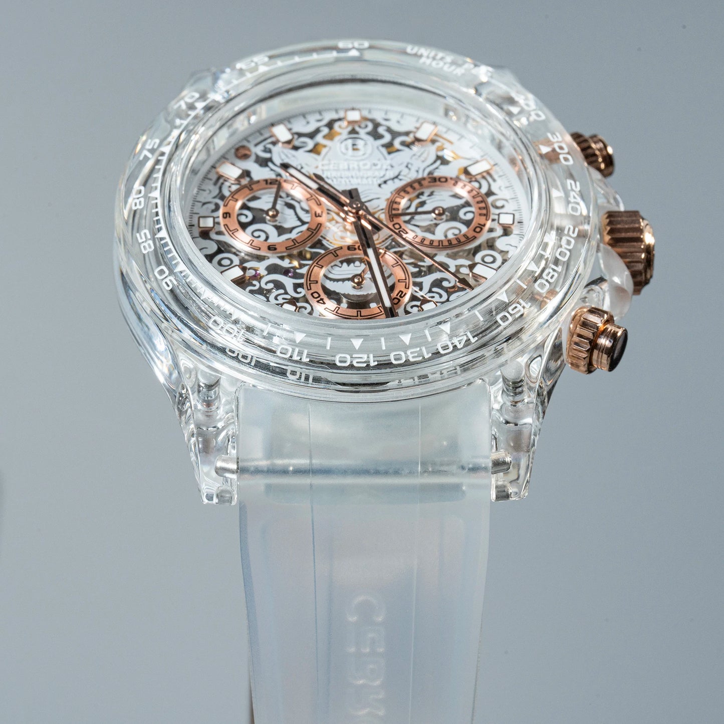 A clear acrylic chronograph watch with a transparent strap and rose gold-toned dials and pushers, displayed on a neutral gray background.