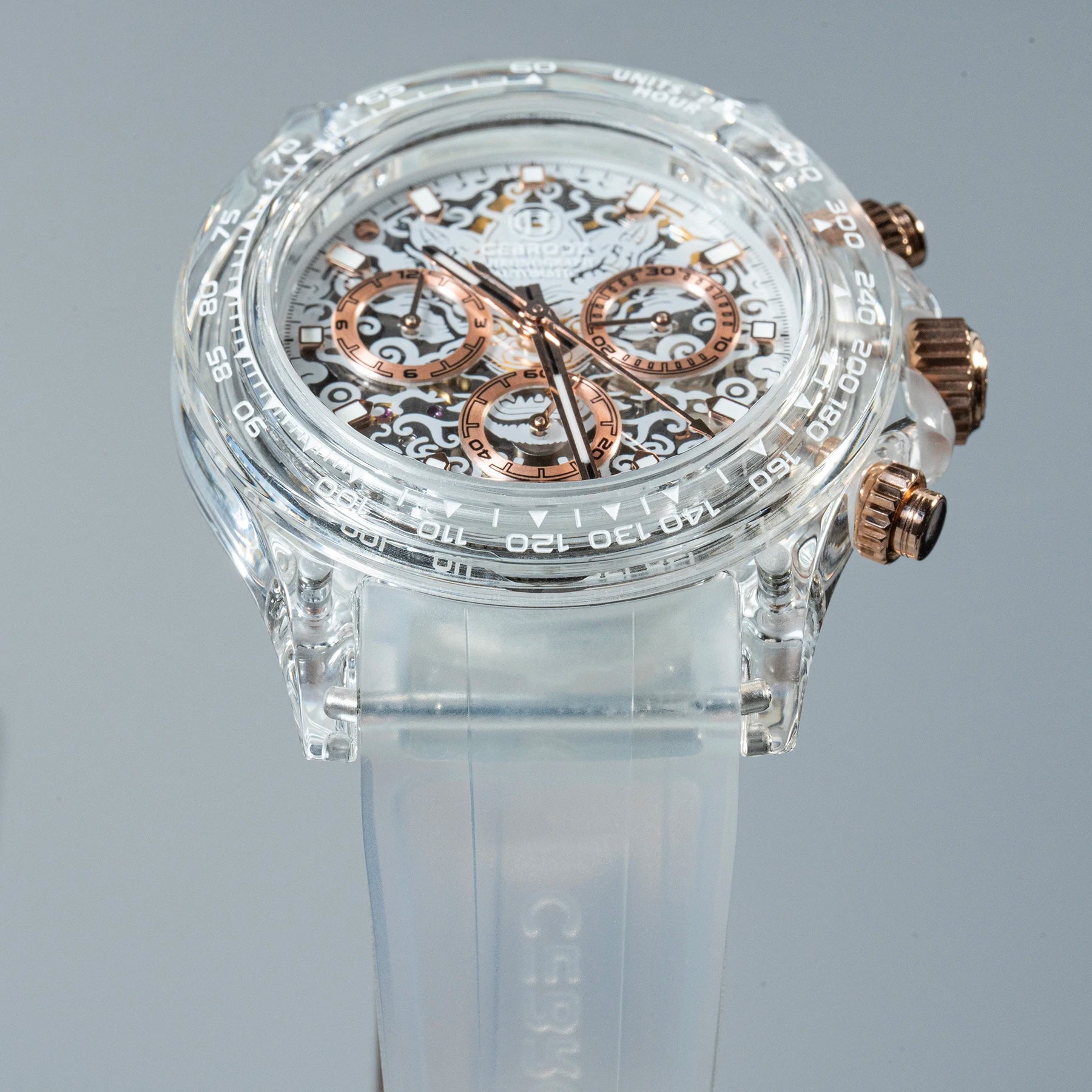 A clear acrylic chronograph watch with a transparent strap and rose gold-toned dials and pushers, displayed on a neutral gray background.