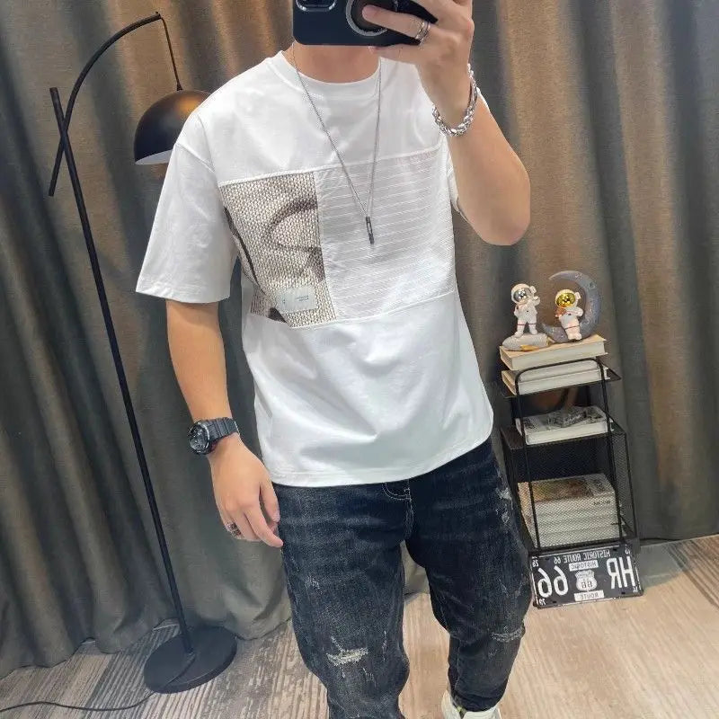 Person wearing a white t-shirt with a patterned design and dark jeans, standing in a room with curtains and a shelf.
