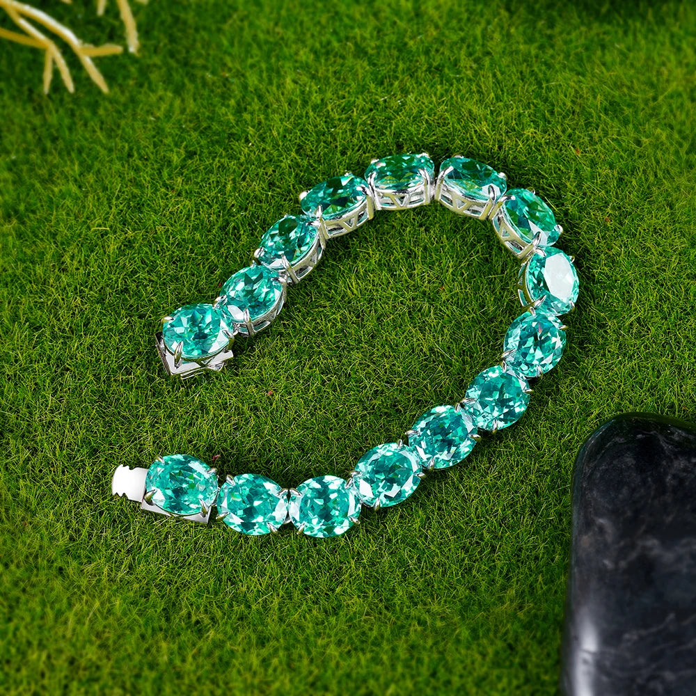 A silver-toned tennis bracelet featuring large oval-cut emerald green gemstones, arranged in a curved shape on a textured green grass background.