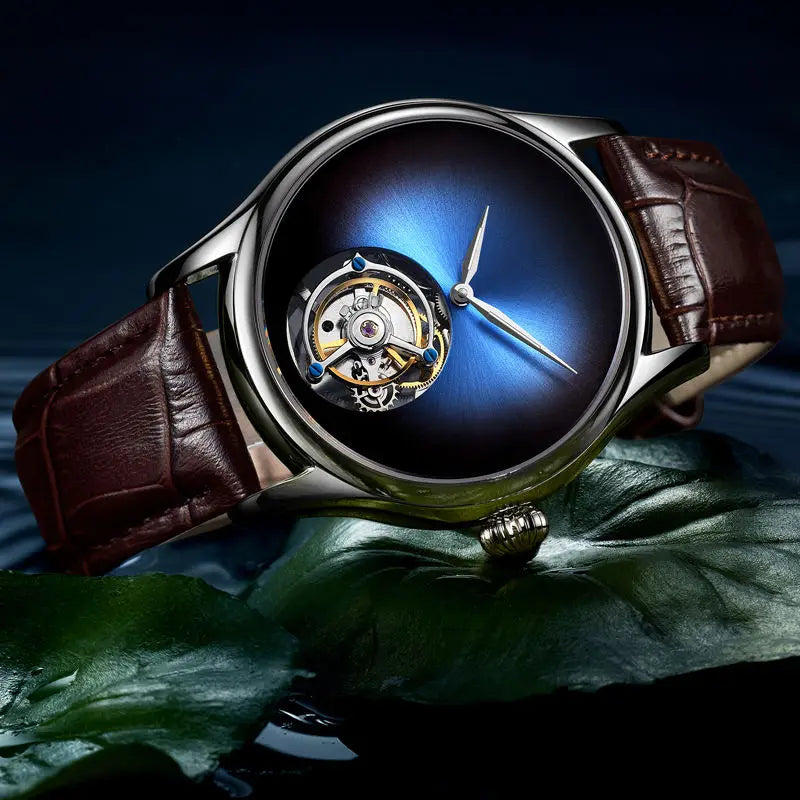 A luxury wristwatch with a dark blue gradient dial, an open-heart tourbillon mechanism, a silver-tone case, and a brown leather strap, resting on a glossy green leaf against a dark background.