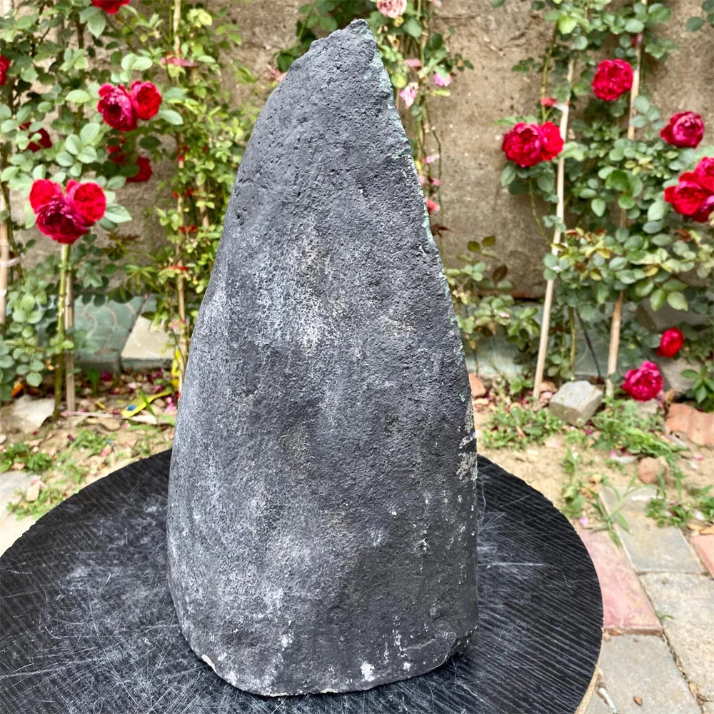 A dark gray textured stone sculpture shaped like a cone or hat, displayed outdoors on a round black table with a garden background featuring red roses.
