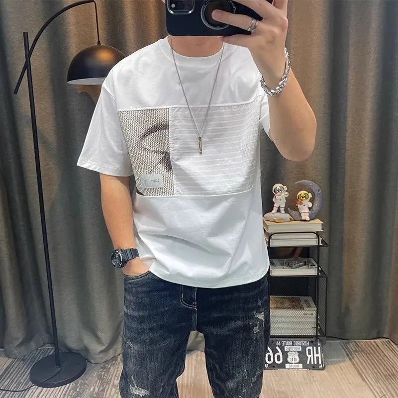 Person wearing a white t-shirt with a patterned pocket and dark jeans, standing in a room with a lamp and shelves.