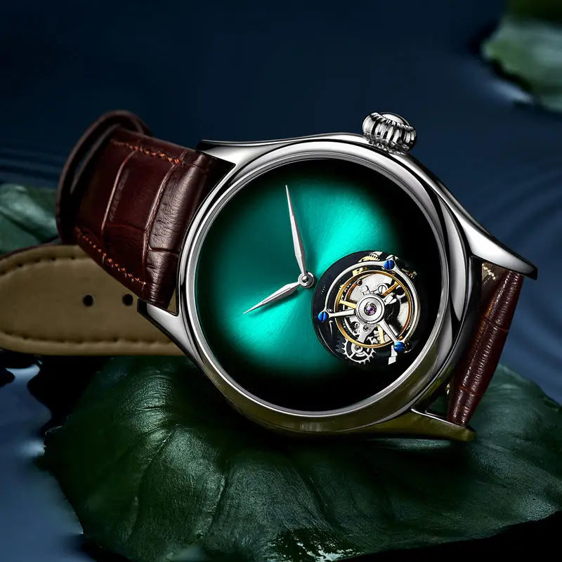 A luxury men's watch with a polished silver-tone case, a green-to-black gradient dial, and a visible mechanical tourbillon at the 6 o'clock position. The watch is fitted with a brown leather strap and is displayed resting on a dark green leaf against a dark blue background.