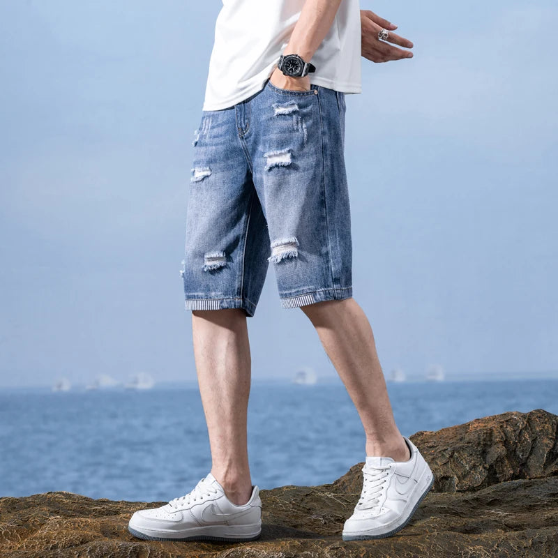 Person wearing ripped blue denim shorts and white sneakers standing on rocks with a blue sky and water background.