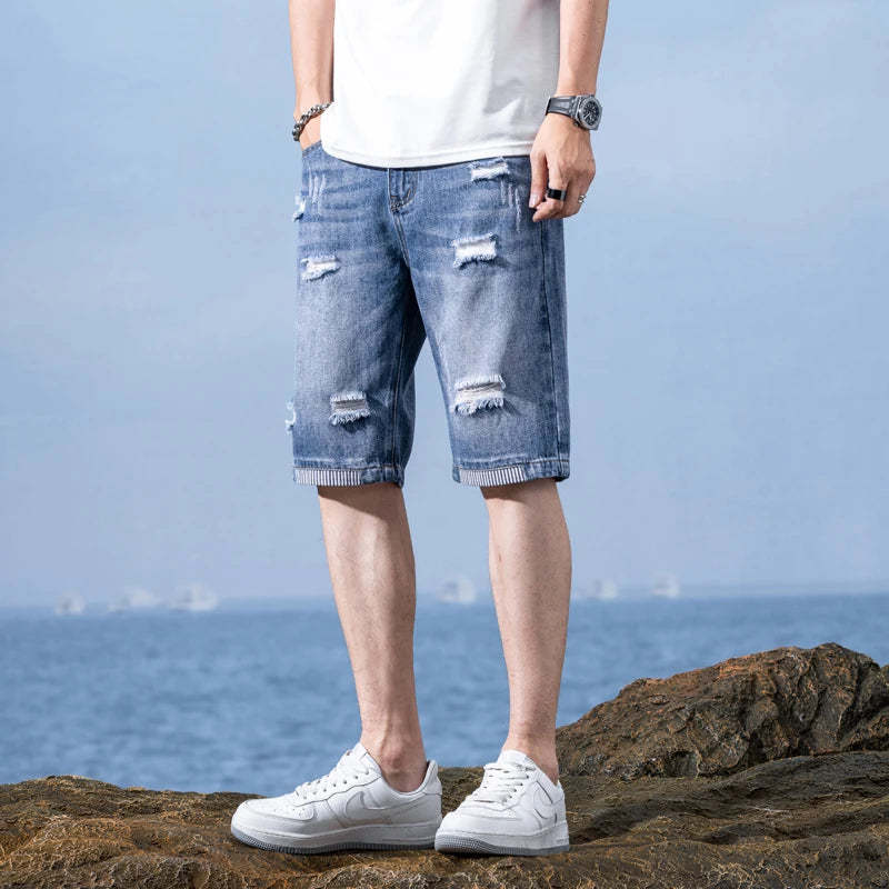 Person wearing ripped blue denim shorts and white sneakers standing on rocks with a blue sky and water background.